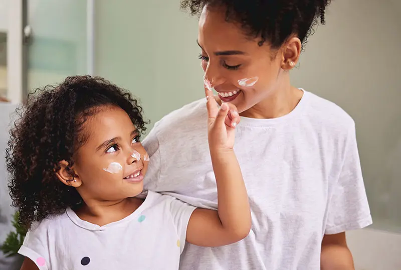 A child and woman with curly hair apply facial cream, smiling at each other.